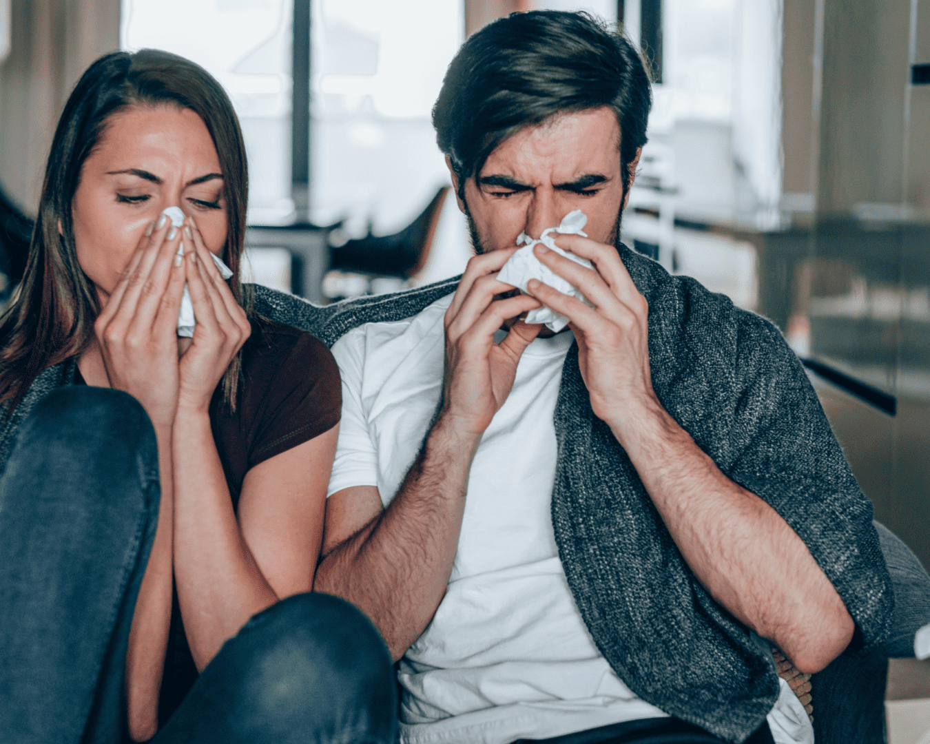 A man and woman sitting on the couch blowing their noses.