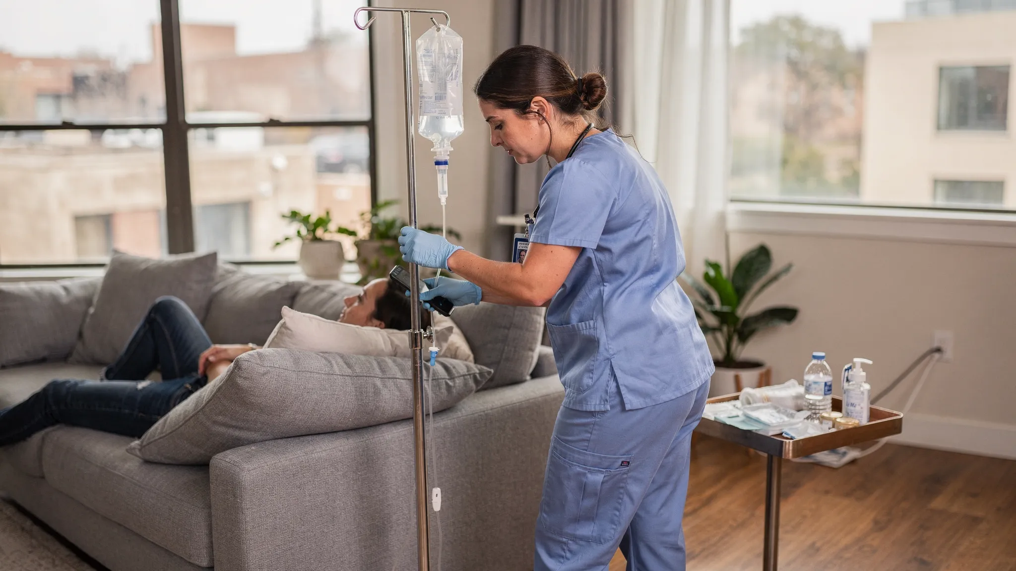 A licensed registered nurse prepares a clear IV hydration bag on a portable stand beside a client relaxing on a sofa in a modern Austin living room, with careful sterile technique and a calm, professional atmosphere.