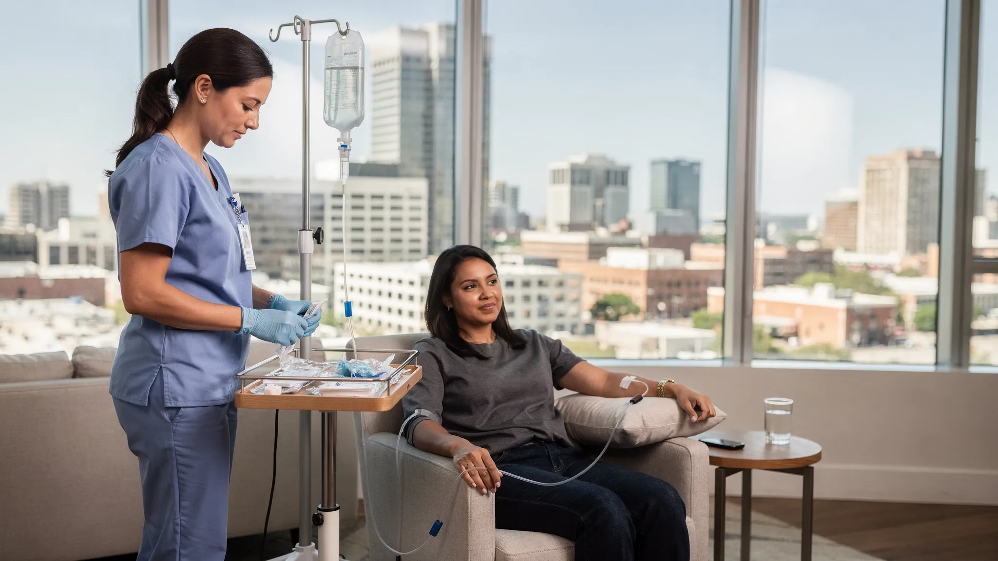 A registered nurse sets up a mobile IV drip for a relaxed client in a modern Austin apartment with floor to ceiling windows and the downtown skyline visible in the background. The nurse wears gloves and uses sterile supplies on a portable tray; an IV bag and tubing hang from a compact stand near a comfortable armchair.